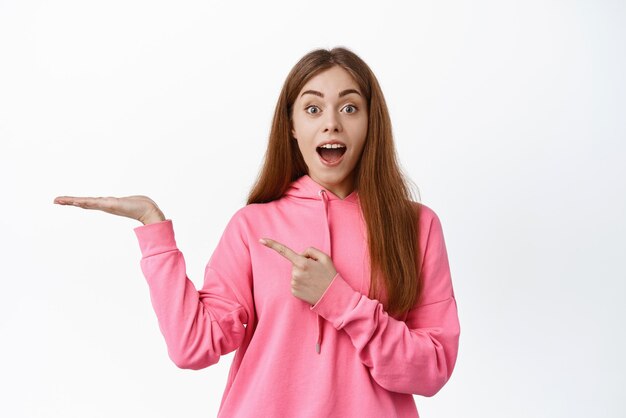 Wow look here Amazed young woman open mouth fascinated pointing at her hand as display object showing your logo standing over white background