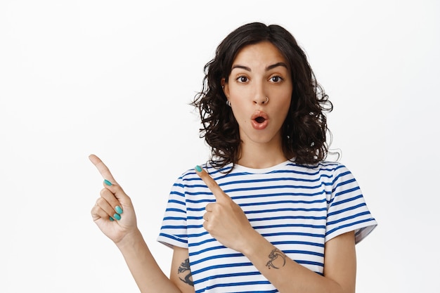 Wow look this way. Amazed brunette woman look impressed, pointing left at logo banner, showing advertisement on empty space, standing white