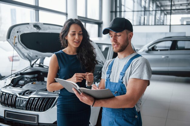 Writing using the pen. Woman in the auto salon with employee in blue uniform taking her repaired car back