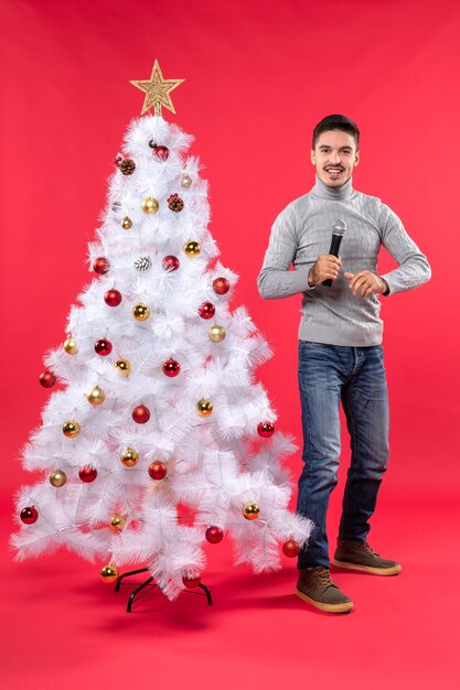 xmas mood with confident guy dressed in jeans standing near decorated Christmas tree holding microphone and speaking happily