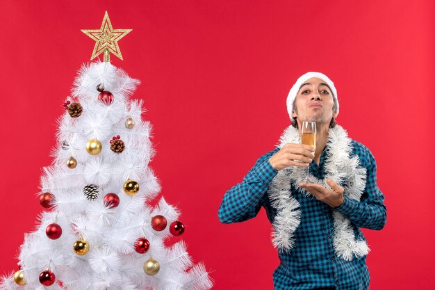 xmas mood with emotional young man with santa claus hat in a blue stripped shirt holding a glass of wine under his chin near Christmas tree