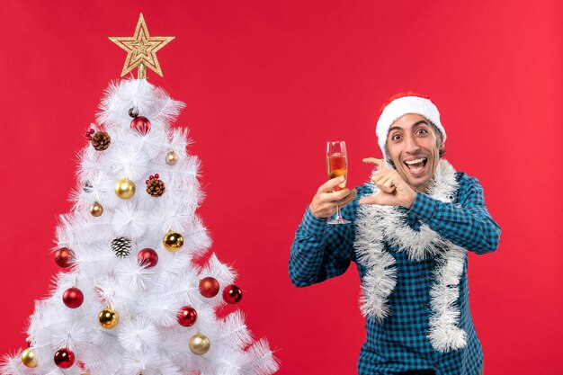 xmas mood with emotional young man with santa claus hat in a blue stripped shirt holding a glass of wine pointing something near Christmas tree