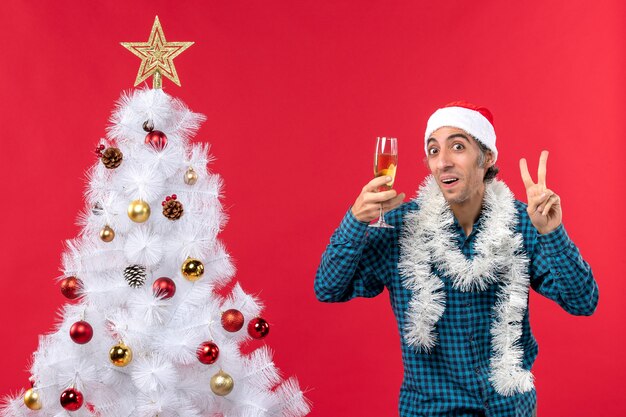 xmas mood with emotional young man with santa claus hat in a blue stripped shirt holding a glass of wine and showing two near Christmas tree