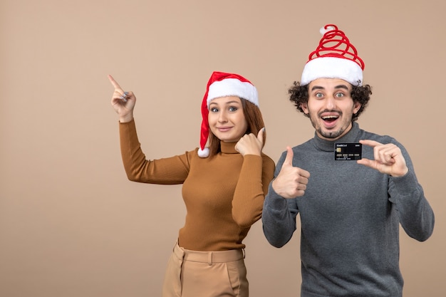 xmas mood with excited satisfied cool couple wearing red santa claus hats guy showing bank card , woman pointing above both doing perfect gesture