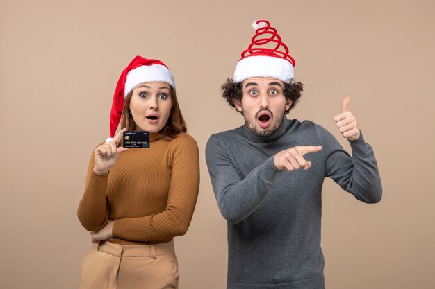 xmas mood with excited satisfied cool couple wearing red santa claus hats woman showing bank card guy pointing something