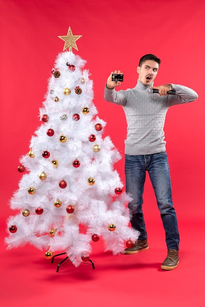 xmas mood with young guy standing near decorated Christmas tree and holding microphone and taking his photo