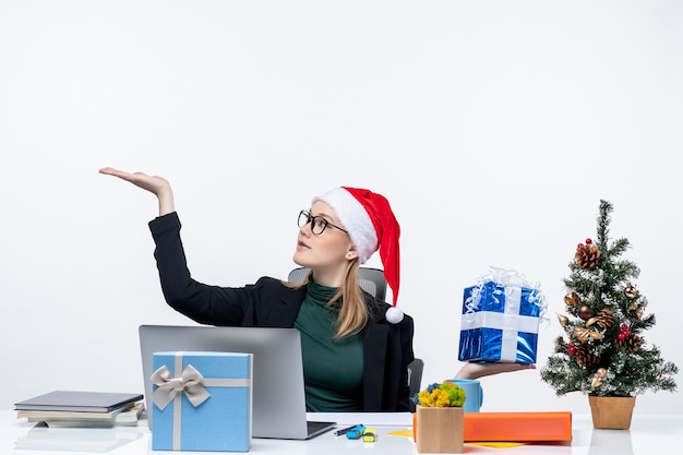 Free photo xmas mood with young woman with santa claus hat and wearing eyeglasses sitting at a table showing gift pointing something on white background