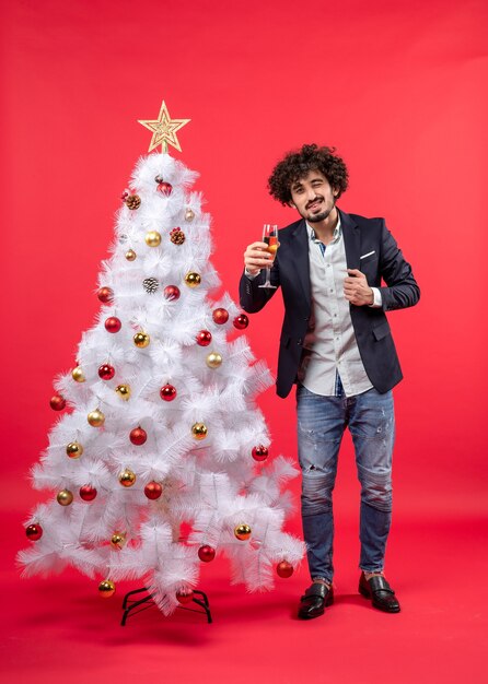 Xsmas celebration with bearded young man with winelooking at camera and standing near Christmas tree on red