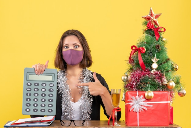 Free photo xsmas mood with concentrated beautiful lady in suit wearing medical mask pointing calculator in the office on yellow