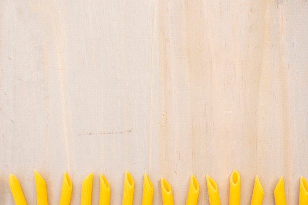 Yellow uncooked penne pasta arranged in row on wooden textured background