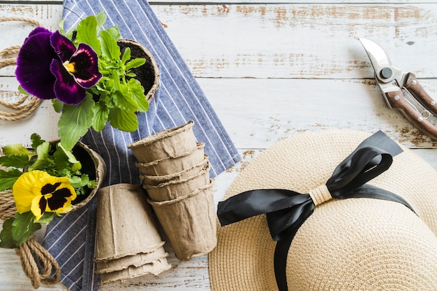 Free photo yellow and violet pansy flowering plant with secateurs; hat; peat pots and napkin on wooden desk