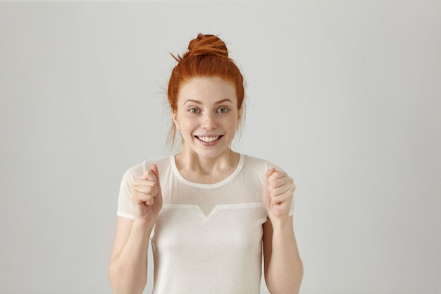 Free photo yes! happy successful young redhead woman winner with hair bun keeping fists clenched while cheering and feeling lucky, looking in excitement and joy, smiling cheerfully