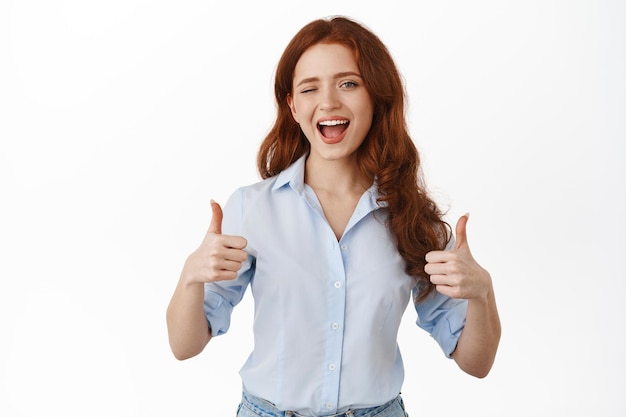 Free Photo yes very good. smiling redhead woman cheering up, winking and showing thumbs up in approval, praise great work, nice job, make compliment, standing in blouse against white background