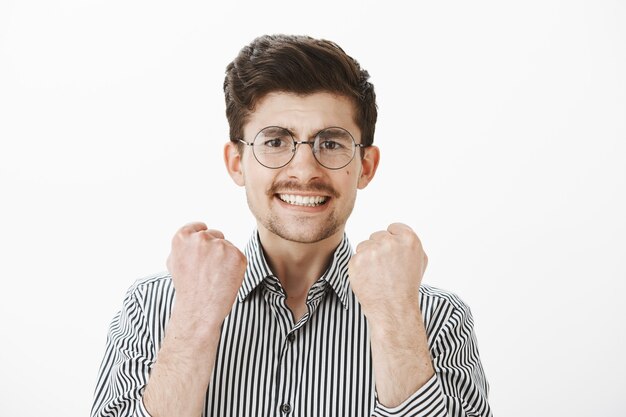 Yes, we finished project in time. Portrait of handsome mature male model in eyewear and striped shirt, raising clenched fists, being confident, celebrating success and victory over gray wall