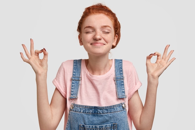 Free photo yoga and meditation concept. calm pleased freckled woman with soft skin, stands with closed eyes spreads hands in zen gesture, wears casual t shirt and denim overalls, isolated over white wall