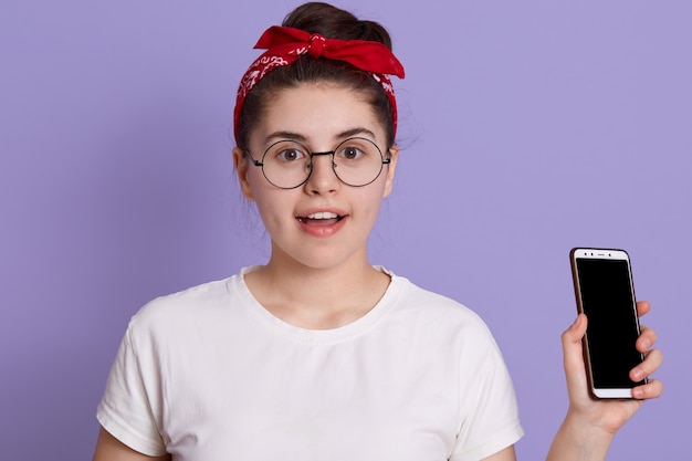 Free photo yong pretty female standing isolated over lilac space  with excitement, holding phone, wearing casual attire and glasses