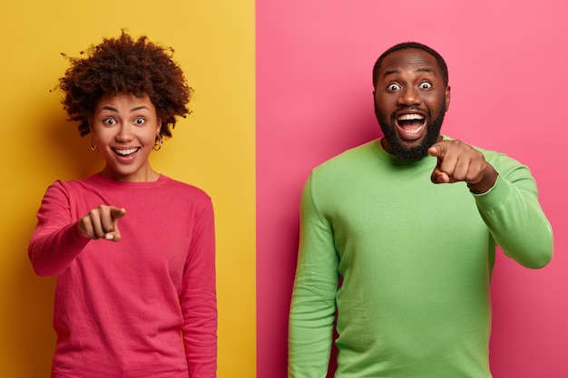 Free photo you are what we need. positive surprised afro american woman and man smile broadly and indicate directly at camera, make choice