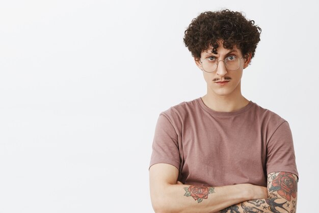 You cannot trick him. Portrait of intense suspicious and doubtful stylish modern hipster guy with fancy moustache tattooed arms and curly dark hair looking from under forehead doubtful in glasses