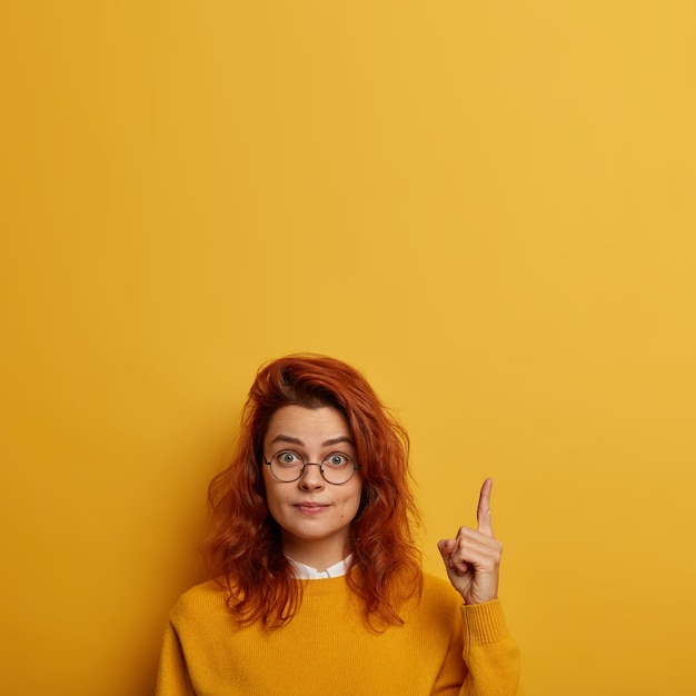 Free Photo you need look. surprised redhead european woman wears round glasses, points above, shows brand new product, advertises copy space