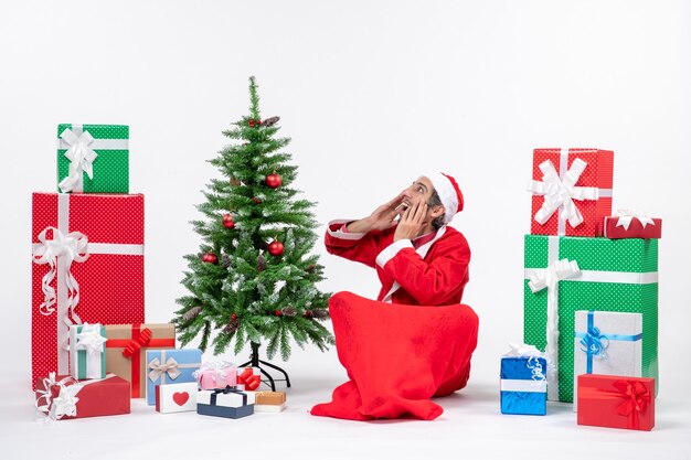 Young adult dressed as Santa claus with gifts and decorated Christmas tree sitting in the ground speaking to someone in surprise