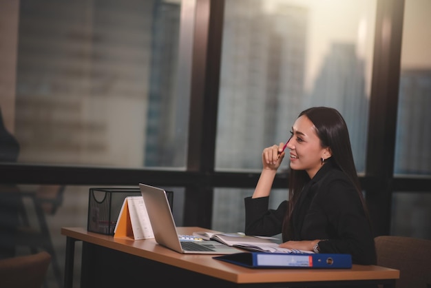 Young adult smart asian business woman in black casual suit using laptop working overtime in urban office