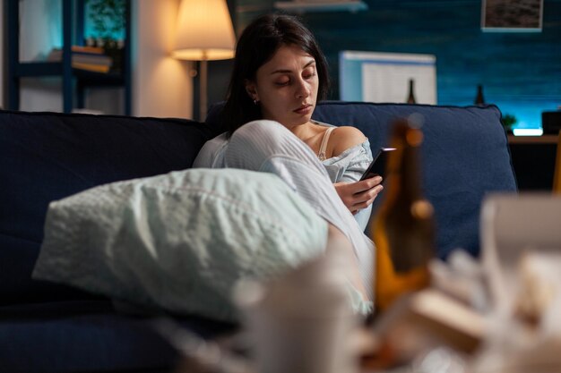 Young adult with anxiety and suicidal thoughts using smartphone to browse internet and search website online. Depressed woman with mental health illness using mobile technology on sofa.