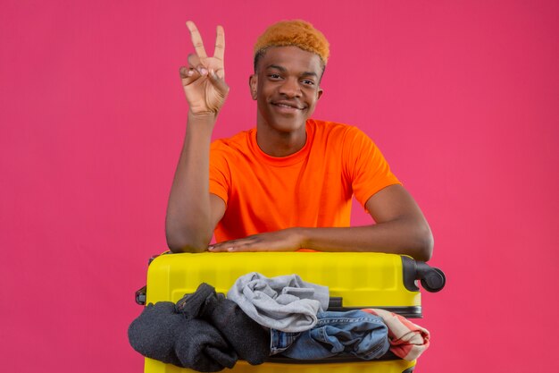 Young african american boy wearing orange t-shirt with travel suitcase full of clothes looking at camera optimistic and cheerful smiling showing number two or victory sign over pink background