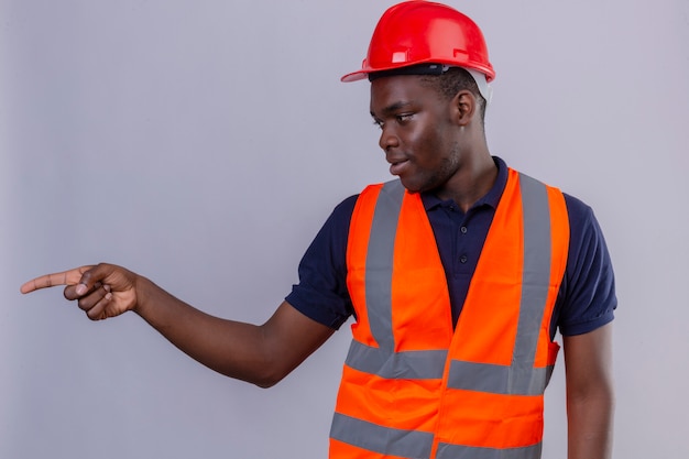 Young african american builder man wearing construction vest and safety helmet looking aside and pointing with finger to the side standing 