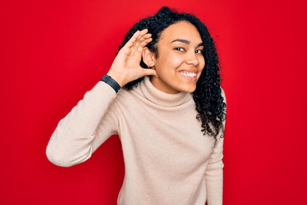 Free Photo young african american curly woman wearing casual turtleneck sweater over red background smiling with hand over ear listening an hearing to rumor or gossip deafness concept