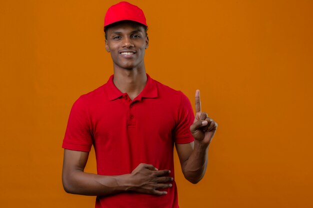 Young african american delivery man wearing red polo shirt and cap standing with smile on face pointing finger up over isolated orange