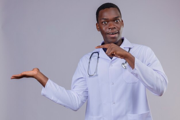 Young african american doctor wearing white coat with stethoscope looking surprised and happy pointing with finger to the side presenting with arm oh his hand