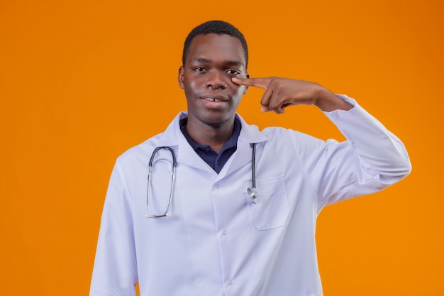 Free photo young african american doctor wearing white coat with stethoscope pointing finger to his eye smiling and waiting