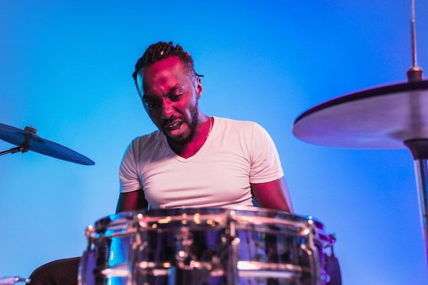 Free Photo young african-american jazz musician or drummer playing drums on blue studio background in trendy neon lights. concept of music, hobby, inspirness. colorful portrait of joyful  artist.