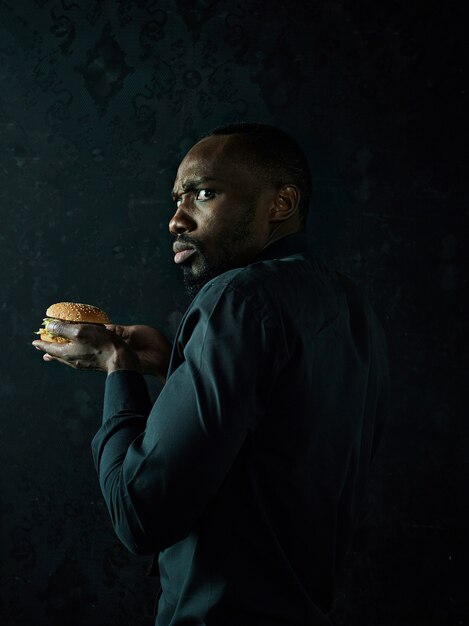 The young african american man eating hamburger and looking away on black studio