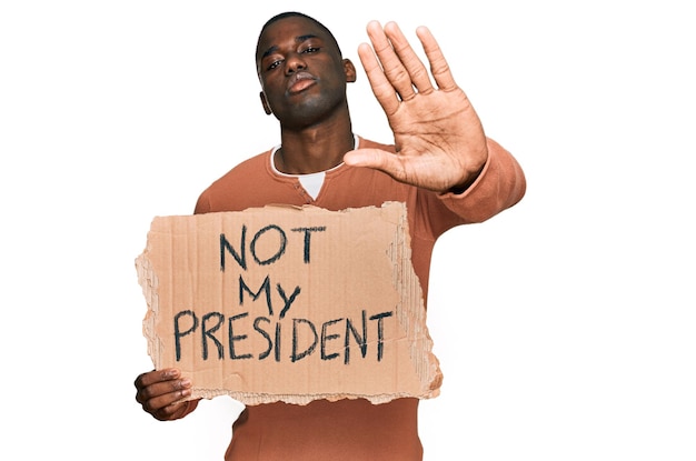 Young african american man holding not my president protest banner with open hand doing stop sign with serious and confident expression defense gesture