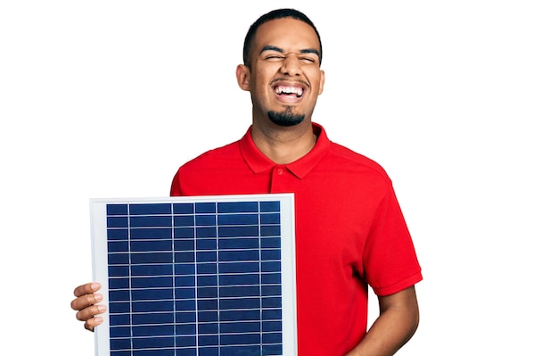 Young african american man holding photovoltaic solar panel celebrating crazy and amazed for success with open eyes screaming excited.