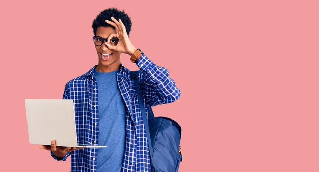 Young african american man holding student backpack using laptop smiling happy doing ok sign with hand on eye looking through fingers