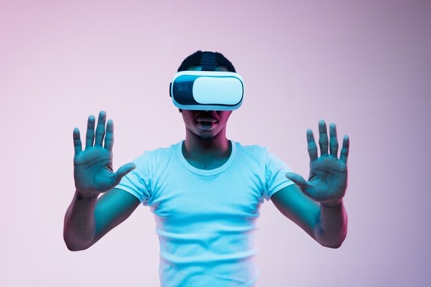 Young african-american man's playing and using VR-glasses in neon light on gradient background