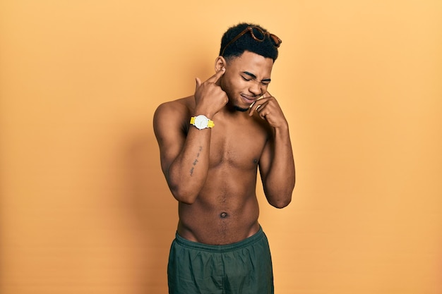 Young african american man wearing swimwear and swimmer glasses covering ears with fingers with annoyed expression for the noise of loud music deaf concept