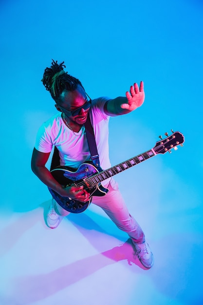 Young african-american musician playing the guitar like a rockstar on blue  background in neon light.