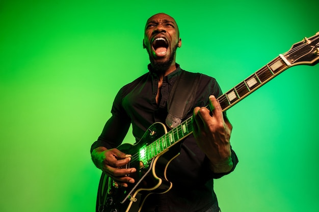 Young african-american musician playing the guitar like a rockstar on gradient green-yellow background.