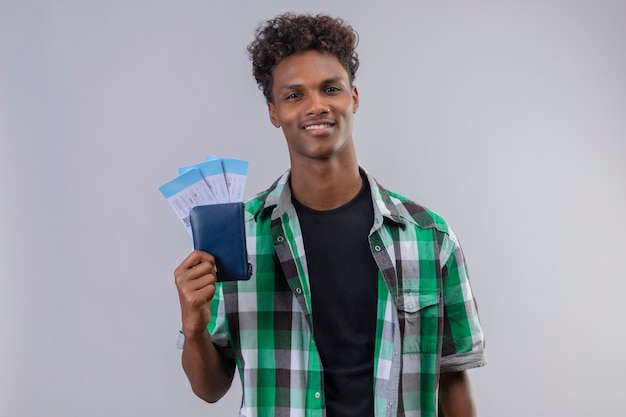 Young african american traveler man holding air tickets smiling cheerfully positive and happy looking at camera standing over white background