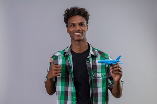 Young african american traveler man holding toy airplane smiling positive and happy showing thumbs up standing over white background