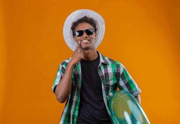 Young african american traveler man in summer hat wearing black sunglasses holding inflatable ring looking at camera with big smile on face pointing with finger to his smile  over ora