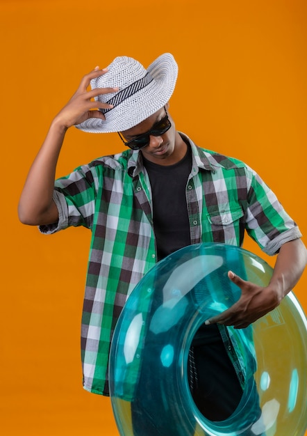 Young african american traveler man in summer hat wearing black sunglasses holding inflatable ring looking down touching his hat