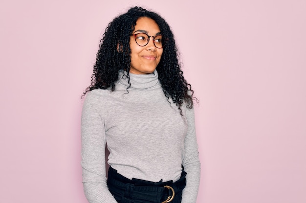 Young african american woman wearing turtleneck sweater and glasses over pink background smiling looking to the side and staring away thinking