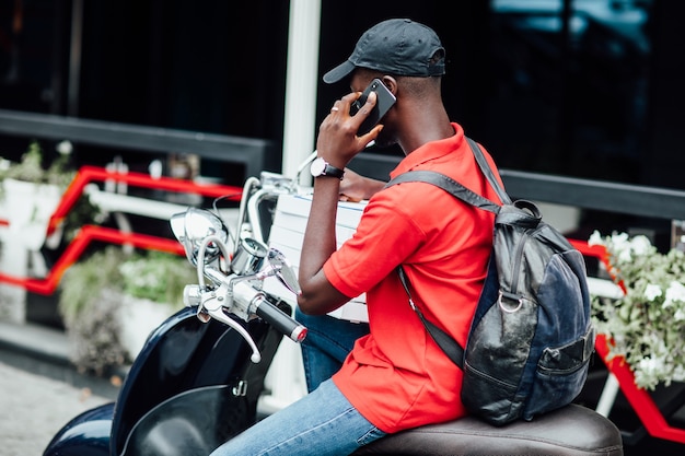 Young African guy accepts the order by phone and write in motorbike holding boxes with pizza