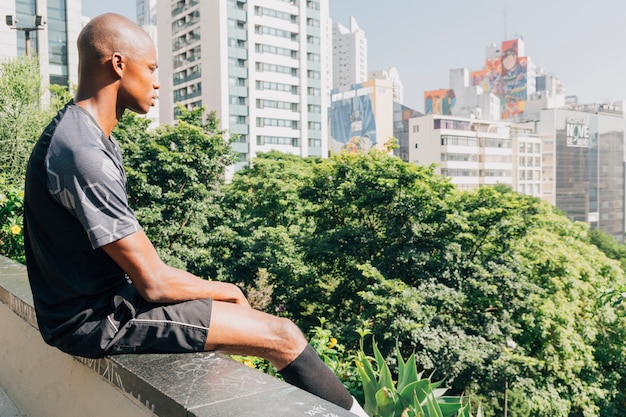 Free Photo young african male athlete sitting on the edge of rooftop overlooking the city