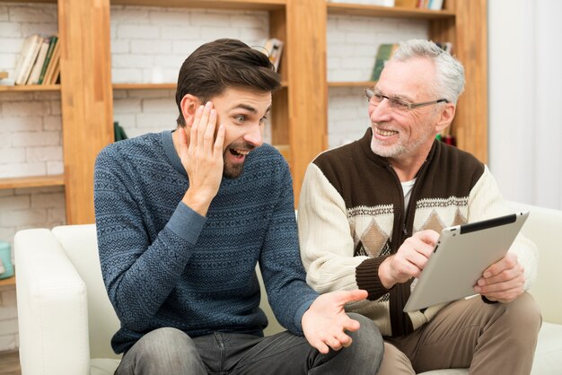 Young amazed guy and aged cheerful man using tablet on settee