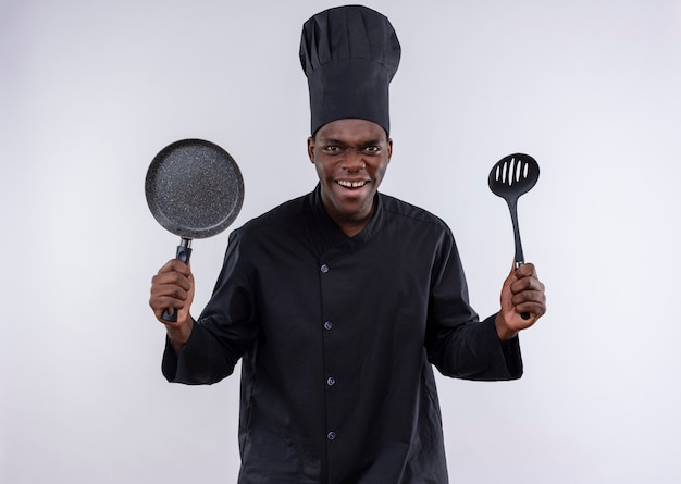 Free photo young annoyed afro-american cook in chef uniform holds frying pan and spatula on white  with copy space